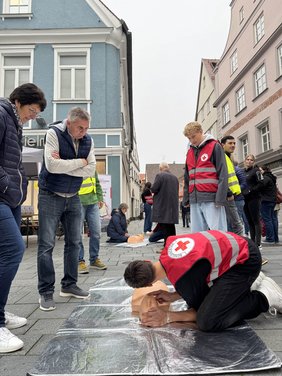 In der Fußgängerzone liegt eine Matte mit Übungspuppe auf dem Boden. Ein Schüler demonstriert Handgriffe an der Puppe, Passanten stehen drumherum und sind aufmerksam dabei.