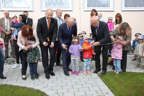 Das Bild zeigt im Vordergrund die Ehrengäste, die mit Unterstützung von Kindern ein Band in Stadtfarben durchschneiden. Rundherum stehen Zuschauer.