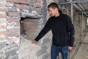 Herr Kögl steht neben einer Schießscharte mit einer breiteren Öffnung nach außen.