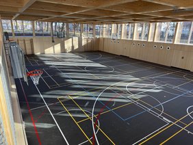 Die leere Halle von der Empore aus. Der dunkle Sportboden ist auffällig, die bunten Linien für die unterschiedlichen Sportarten sind deutlich zu erkennen. 