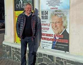 Zu sehen ist Otfried Richter vor einer Hauswand. An der Wand hängt ein Plakat, auf dem groß ein Porträt von Otfried Richter zu sehen ist und im ukrainischen Text ein Konzert mit Chor und Gastdirigent angekündigt wird.