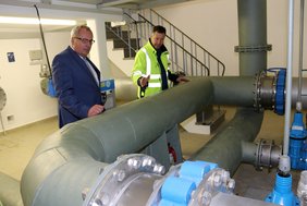 Die beiden Männer stehen hinter großen Rohrleitungen, die einen Durchmesser von ca. 40 bis 50 cm haben.