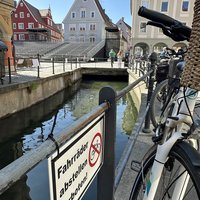 Im Vordergrund ein Schild "Fahrräder abstellen verboten", das am Geländer zum Stadtbach befestigt ist. Direkt daneben ein Fahrrad. 