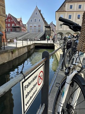 Im Vordergrund ein Schild "Fahrräder abstellen verboten", das am Geländer zum Stadtbach befestigt ist. Direkt daneben ein Fahrrad. 