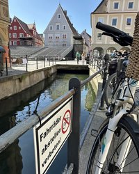 Im Vordergrund ein Schild "Fahrräder abstellen verboten", das am Geländer zum Stadtbach befestigt ist. Direkt daneben ein Fahrrad. 