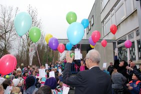 Luftballons steigen in den wolkenverhangenen Himmel auf. Unten sind Kinder zu sehen und OB Holzinger.