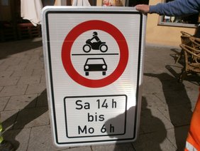 Dargestellt ist ein Verkehrsschild mit einem roten Kreis, der ein Verbot für Motorräder und PKW anzeigt. Dazu die durchfahrtsgesperrten Zeiten: Samstag 14 Uhr bis Montag 6 Uhr.