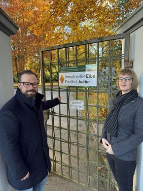 Die beiden Personen stehen an der Gittertür zum Friedhof, an der das Schild montiert ist. Das Schild hat die Aufschrift: "Immaterielles Erbe Friedhofskultur". 