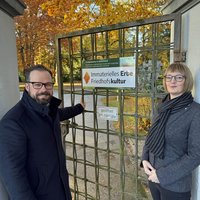 Die beiden Personen stehen an der Gittertür zum Friedhof, an der das Schild montiert ist. Das Schild hat die Aufschrift: "Immaterielles Erbe Friedhofskultur". 
