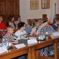 Eine Reihe von Kindern sitzen an den Tischen im Sitzungssaal. Ein Mädchen meldet sich engagiert. Im Hintergrund sitzen zuschauende Eltern und Lehrer. 