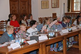 Eine Reihe von Kindern sitzen an den Tischen im Sitzungssaal. Ein Mädchen meldet sich engagiert. Im Hintergrund sitzen zuschauende Eltern und Lehrer. 