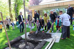 zu sehen sind die vielen Erwachsenen und Kinder mit Schaufeln, die gemeinsam Erde in ein Pflanzloch schaufeln.